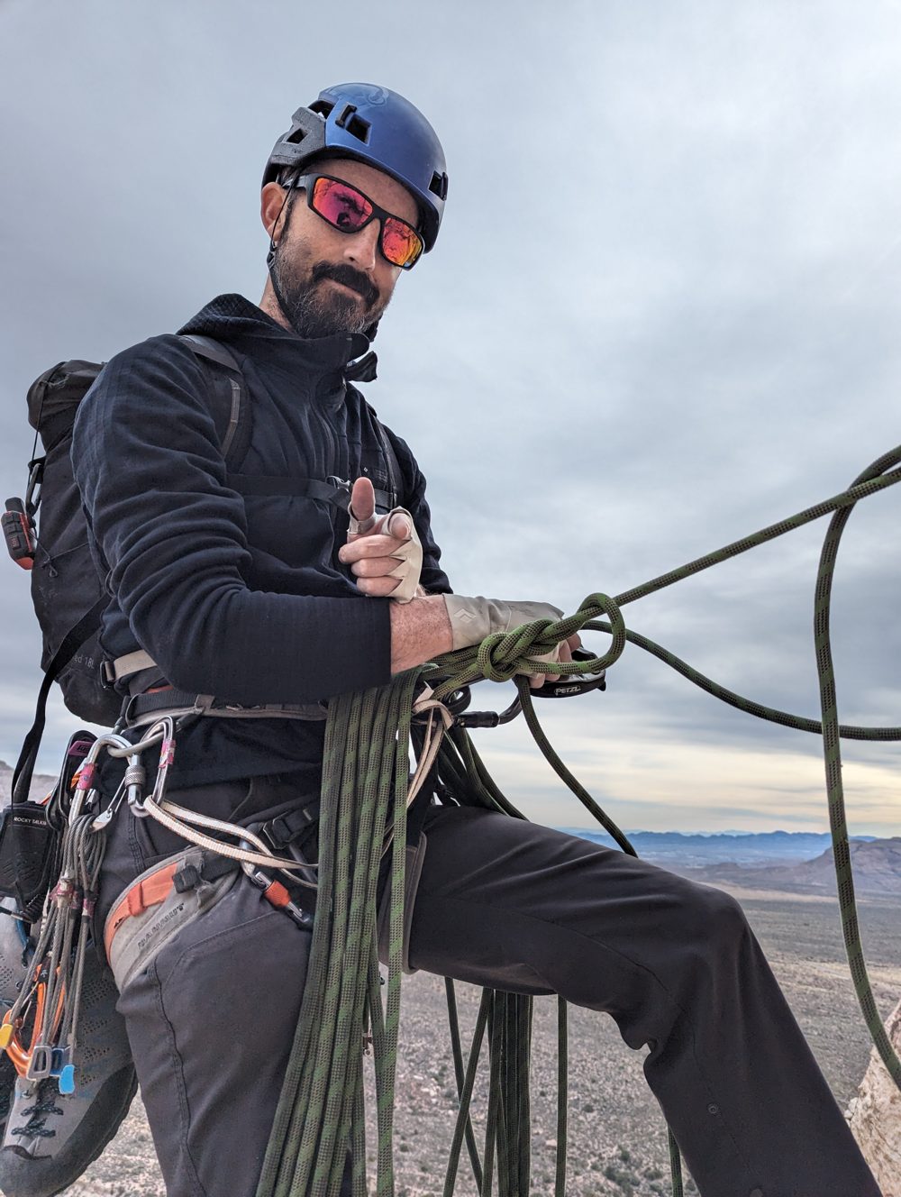 Colin in a helmet and sunglasses, high on a cliff with a coiled rope and a desert valley behind him