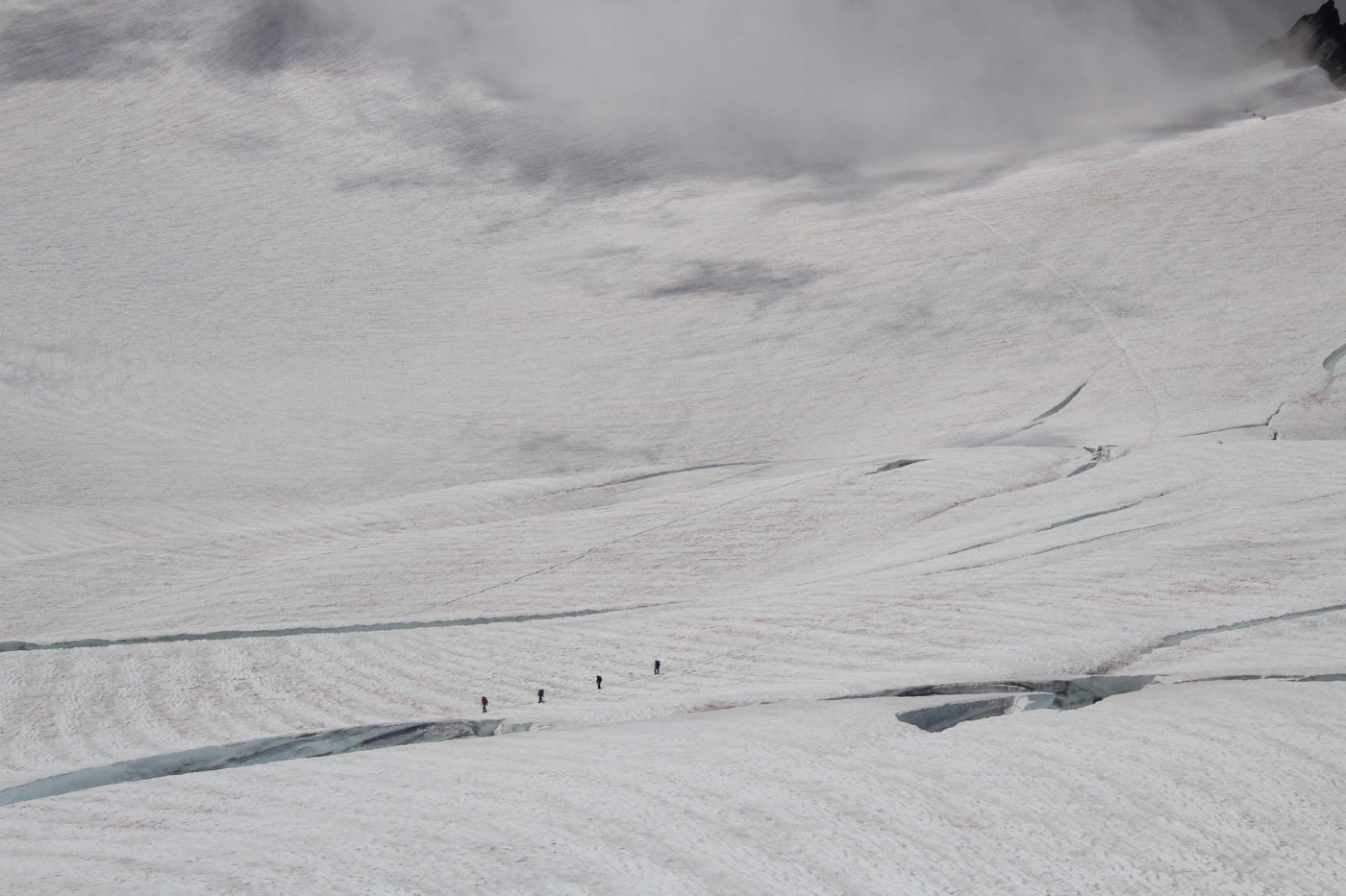 A roped team of climbers crossing a snowfield below cloud-covered peaks, crevasses visible nearby