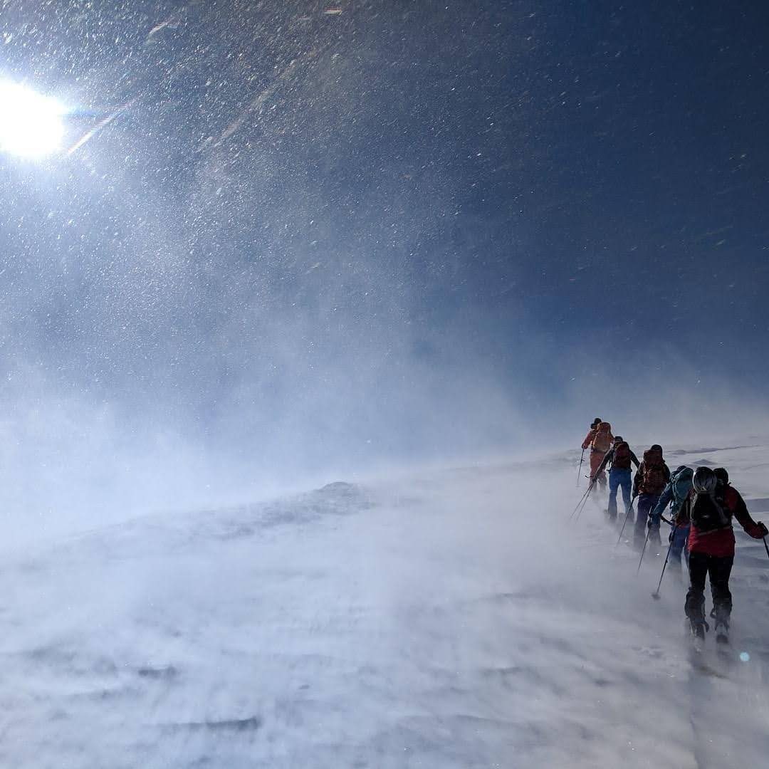 Colin and a line of backcountry skiers ascending a wind-blown ridge in bright sun and blowing snow